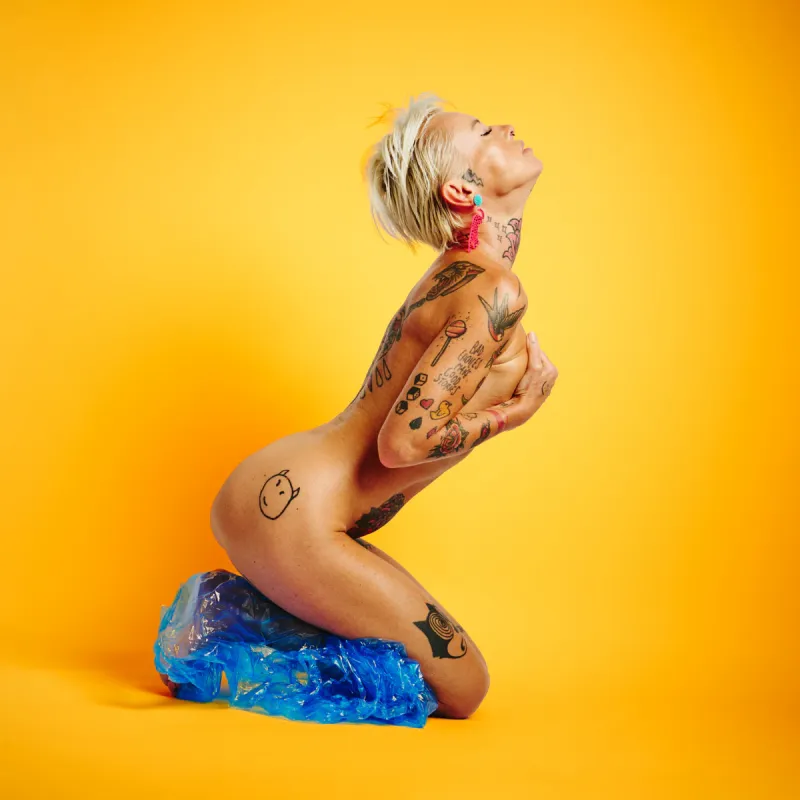 Lu Epic nude wearing blue transparent plastic pants against a strong yellow background in the Colors series