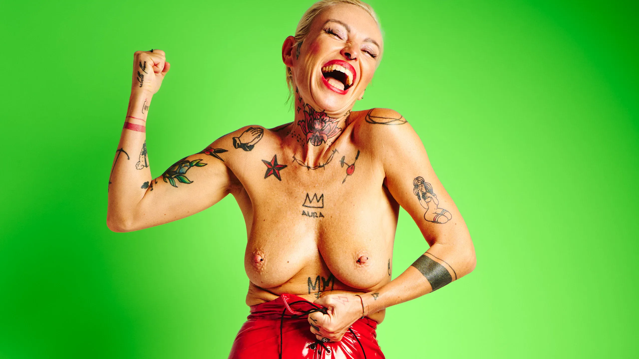 Lu Epic in an energetic topless art series wearing red latex shorts and heels against green