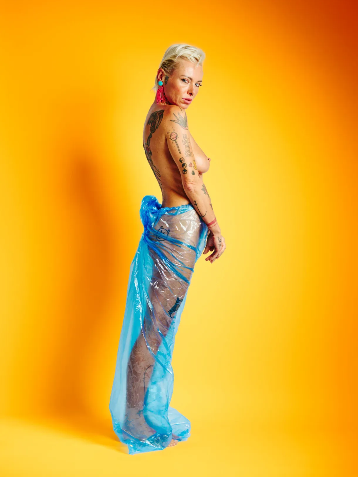 Lu Epic nude wearing blue transparent plastic pants against a strong yellow background in the Colors series