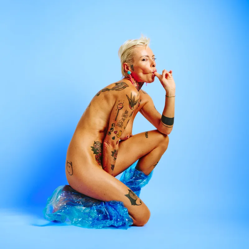 Explicit art nude portrait of Lu Epic in blue plastic pants with vivid yellow backdrop